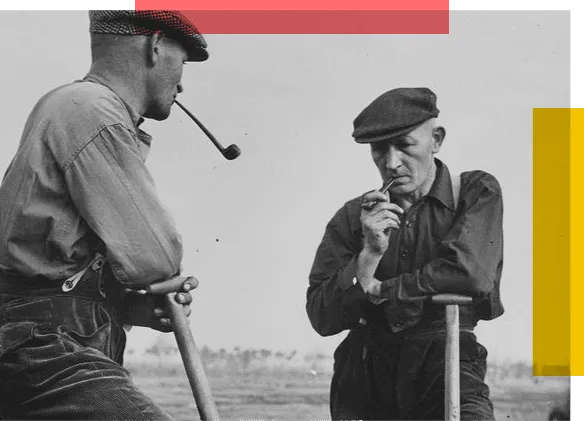 Two men with pipes stand leaning on long-handled tools looking at each other.